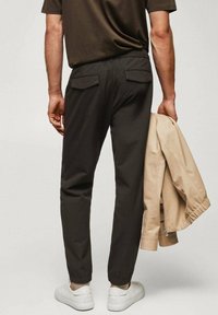 Ciemnoszare spodnie z elastycznymi ściągaczami i tylnymi kieszeniami, zestawione z jasnobeżową kurtką trzymaną w ręce. Całość dopełniają białe sneakersy.