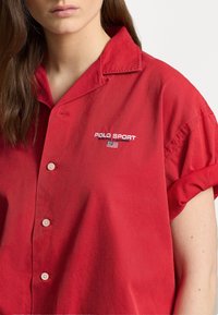 Red short-sleeve shirt made of cotton with a button-up design. Features embroidered "POLO SPORT" logo and two buttons on the front.