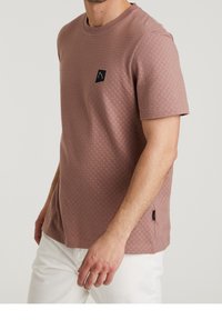 Mężczyzna w fakturowanej, brudnoróżowej koszuli z krótkim rękawem oraz białych spodniach, stojący z ręką swobodnie opartą na biodrze na jednolitym tle.