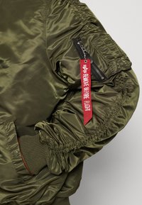 Olive green bomber jacket with shiny, smooth material. Features ribbed cuffs, a zippered pocket, and a red "Remove Before Flight" tag.