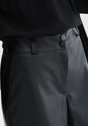 Schwarze Kunstlederhosen mit glatter Textur, hoher Taille und zwei runden schwarzen Knöpfen am Verschluss. Mit genähten Details.