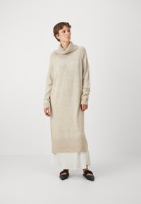Abito in maglia beige con scollo a bottleneck e maniche lunghe, con polsini e orlo a coste, indossato sopra un abito sottoveste bianco. Texture liscia.