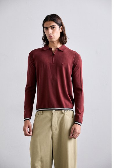 Bordeaux farvet langærmet polo med lynlås krave, ribbet tekstur og et lille logo på brystet. Båret sammen med lyse beige bukser.