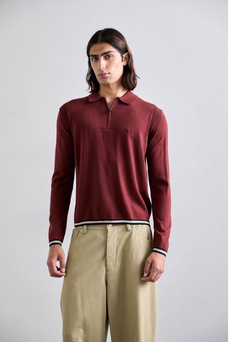Polo a maniche lunghe di color marsala con colletto zip, texture a coste e un piccolo logo sul petto. Abbinato a pantaloni beige chiaro.