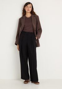 Csokoládébarna zakó egy passzos, hozzáillő ing felett, magas derekú, fekete, plissézett nadrággal és barna cipővel párosítva.