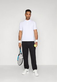 Λευκό μπλουζάκι τένις με κοντά μανίκια και λαιμόκοψη henley, συνδυασμένο με μαύρο παντελόνι. Το άτομο κρατάει ρακέτα και μπάλα τένις.