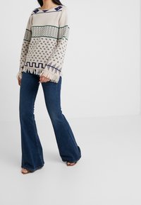 Pull en maille présentant des motifs géométriques en crème, marine et vert avec un ourlet à franges ; porté avec un jean évasé bleu foncé.