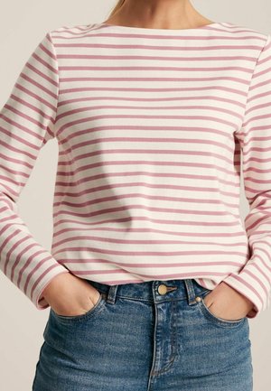 Kobieta w białej, długorękawowej koszulce w różowe poziome paski, włożonej do wysokich, niebieskich dżinsów, z rękami w kieszeniach.