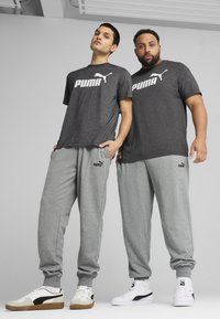 Divi vīrieši, tērpti saskaņotās pelēkajās Puma T-kreklos ar baltām emblēmām, kopā ar pelēkām sporta biksēm un Puma sporta apaviem melnbaltā krāsā.