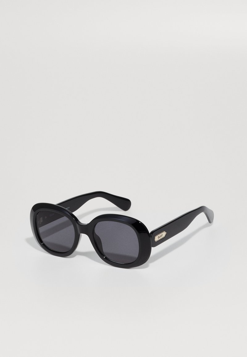 Occhiali da sole oversize neri a forma ovale con montatura spessa e lenti scure, caratterizzati da un piccolo logo oro Quay sul braccio.