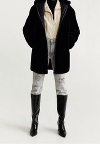 Zwarte jas van nepbont met capuchon, crèmekleurige kabelgebreide trui, verknipte grijze spijkerbroek en zwarte kniehoge puntige laarzen.