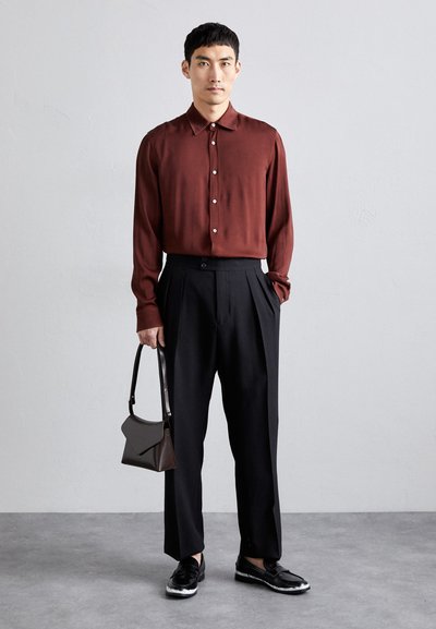 Långärmad vinröd skjorta, svarta plisserade byxor och svarta loafers. Håller en liten brun handväska. Enkel, modern design med knappar.