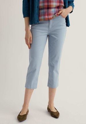 Naine, kes kannab helerohelisi lühemaid pükse, mitmevärvilist ruudulist särki, sinist kardigani ja pruune teravate ninadega kingi, seisab ühe käega taskus.