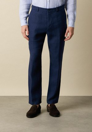 Man draagt marineblauwe op maat gemaakte broek, lichtblauw gestreept overhemd in de broek gestopt en donkerbruine suède loafers, staand op een grijze vloer.