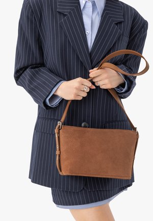 Naine kannab mereväe triibulist ülikonda ja hoiab pruuni seemsnahast käekotti ühe õlarihmaga, taust on valge.