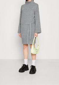 Robe en maille grise avec col roulé, ceinture et larges manches ; portée avec des chaussures noires épaisses et un sac vert clair orné d'un accent décoratif.