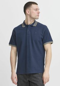 Marinblå polo skjorta med en texturerad yta, med en tvåknappsknäppning och kontrasterande krage och ärmslut i beige ränder.