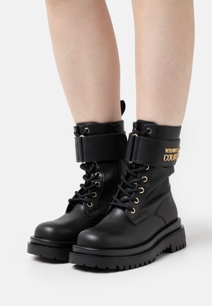 Bottes de combat noires à lacets avec semelles épaisses et sangles en cuir ornées de ferrures dorées, portées sur des jambes nues sur un fond blanc.