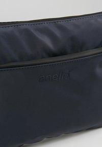 Borsa in tessuto blu navy con superficie liscia, chiusura a zip e una leggera lucentezza. Dotata di tasca frontale e dettaglio del logo in rilievo.