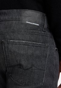 Schwarze Denim-Jeans mit einem Leder-Label-Patch am Bund, hinteren Taschen mit dezenten Nahtdetails und einer strukturierten Stoffoberfläche.