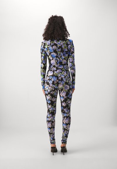 Floral jumpsuit in black, featuring blue and green floral patterns. Long sleeves and form-fitting design, with high-heeled shoes.