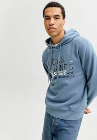 Μπλε φούτερ με κουκούλα από απαλό ύφασμα, με εμπρός εκτύπωση με το λογότυπο "Jack & Jones", κορδόνια και ριμπ τελειώματα στα μανίκια. Φορεμένο με μπλε τζιν.