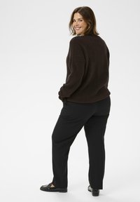 Nő, aki a vállán át mosolyog, sötétbarna bolyhos pulóvert, fekete nadrágot és fekete mokaszin cipőt visel, fehér háttér előtt áll.