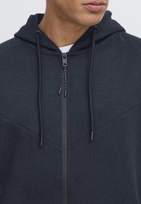 Tamno plava zip-up dukserica s teksturiranim završetkom, s kapuljačom s vezicom i kontrastnim svjetlijim panelima sprijeda.