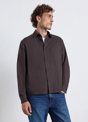 Mann mit lockigem Haar, der ein dunkelbraunes Hemd mit Knöpfen und blaue Jeans trägt, schaut zur Seite vor einem schlichten hellen Hintergrund.
