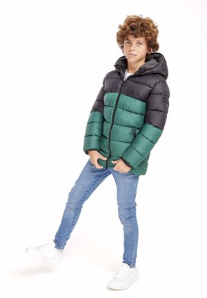 Gewatteerde jas met een zwart bovenstuk en een groene ondersectie, voorzien van een ritssluiting aan de voorkant, een capuchon en zijzakken. Draagt over blauwe spijkerbroeken en witte sneakers.