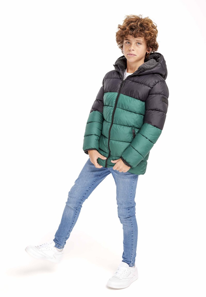 Vadderad jacka med svart överdel och grön nederdel, med dragkedja fram, huva och sidofickor. Bärs över blå jeans och vita sneakers.