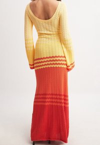 Robe maxi en tricot présentant un dégradé du jaune au rouge, avec un dos nu et des manches longues, arborant un motif ondulé sur toute la surface.
