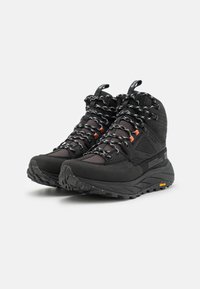 Scarpe da trekking nere con pannelli in mesh, lacci riflettenti e dettagli arancioni. Suole in gomma testurizzata con logo Vibram per una buona aderenza. Design alta.
