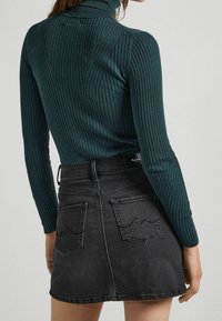 Frau, die einen dunkelgrünen, gerippten Rollkragenpullover trägt, in einen hoch taillierten schwarzen Jeansrock mit Gesäßtaschen und dezenter Stickerei gesteckt.