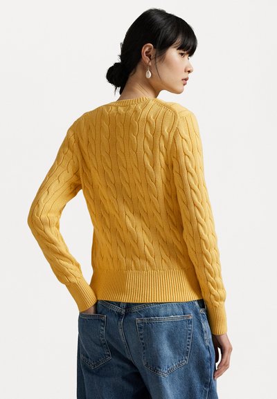 Gelber Kabelstrickpullover mit gerippten Bündchen und Saum, mit einem Design auf der Rückseite. Kombiniert mit lässigen blauen Jeans.