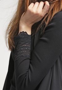 Mustapaita, jossa on pitkät hihat, ribbitekstuuri ja pitsikoristeet hihojen suissa. Hihojen suut ovat yksityiskohtaisia pitsiyksityiskohtia, joissa on aaltoileva reuna.