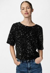 Svart paljett-topp med løs passform, rund halsutskjæring og korte ermer. Teksturert overflate som reflekterer lys, kombinert med blå denimjeans.