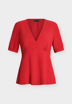 Donna che indossa una camicetta rossa con scollo a V, jeans scuri a gamba larga con risvolto, sandali beige con tacco, che tiene una piccola borsa bianca, in piedi su sfondo bianco.