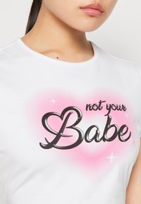 Vit t-shirt med kort ärm som har stor svart text "not your Babe" på en rosa hjärtbakgrund med glittrande accenter. Slät bomullstextur.
