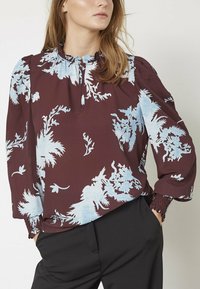 Bordeaux skjorte med lysblå blomsterprint, rynket halsudskæring og puffede ærmer; stoffet har en glat tekstur. Stylet med sorte bukser.