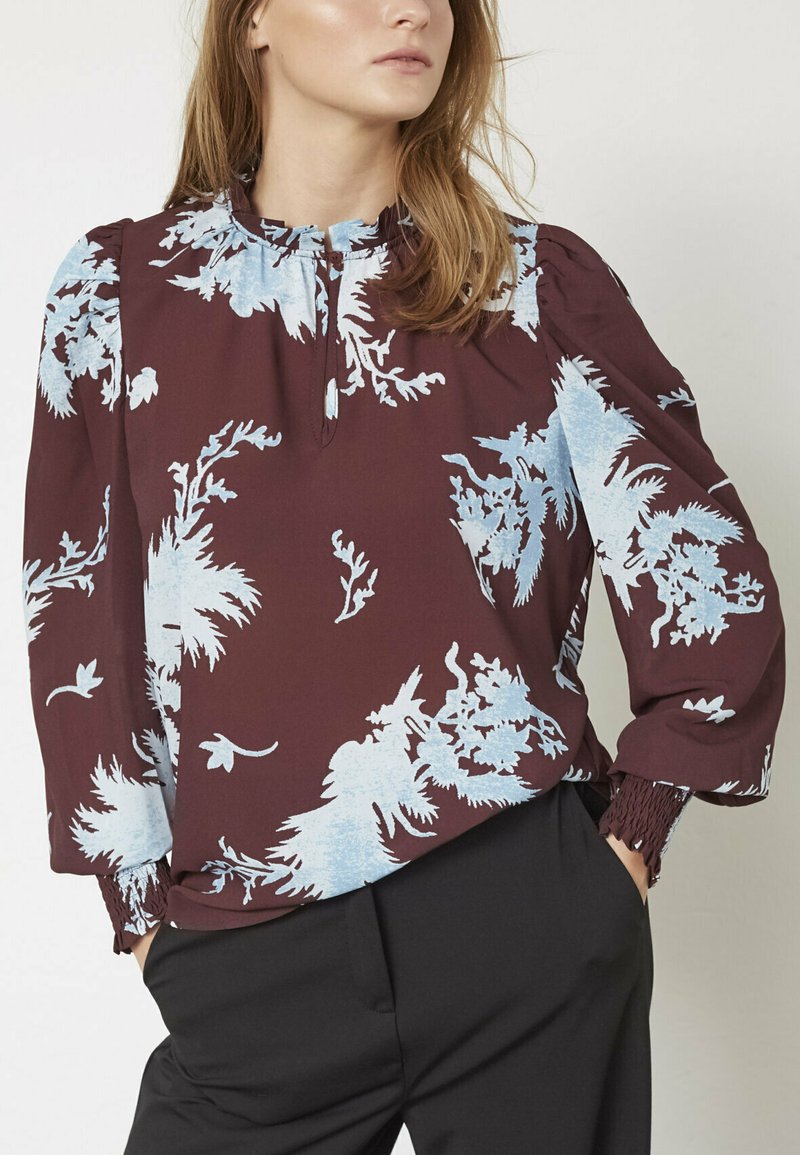 Bordeaux skjorte med lysblå blomsterprint, rynket halsudskæring og puffede ærmer; stoffet har en glat tekstur. Stylet med sorte bukser.