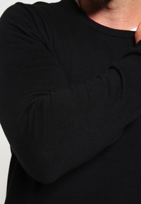 Zwart gebreid langemouwen top met een ronde hals. Voorzien van ribgebreide manchetten en een gladde textuur. Close-up van de mouw en arm.