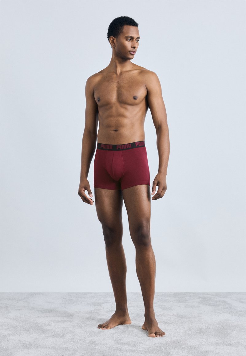 Bordo bokseris ar melnu elastīgu jostasvietu, uz kuras redzams "PUMA" logotips, parādīts stāvoša vīrieša modelī uz vienkārša fona.