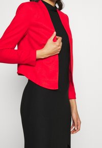 Kvinna som justerar en figursydd, ljusröd kort jacka över en långärmad svart klänning mot en enkel vit bakgrund.