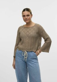 Pullover lavorato a maglia color marrone chiaro con un motivo a diamante, scollo a barca e maniche a campana. Abbinato a jeans larghi di colore blu chiaro.