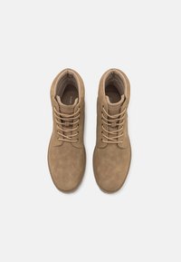 Botas de ante marrón hasta el tobillo con punta redonda, diseño de cordones, cuello acolchado y superficie texturizada. Marca mínima visible en el interior.