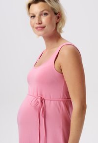 Roze zwangerschapsjurk met een aansluitende bovenkant, verstelbare strik in de taille en brede schouderbanden. Glad, zacht materiaal dat comfort benadrukt.