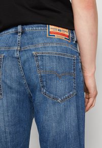 Jeans de mezclilla azules con un diseño clásico de cinco bolsillos, cintura de media altura y una prominente etiqueta de marca en la parte trasera. Detalles de costura intrincados visibles.