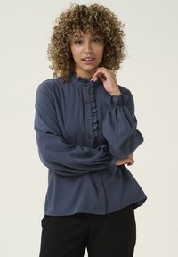 Blouse bleu marine avec col à volants, fermeture par bouton et manches ballon. Fabriquée en tissu lisse avec une coupe décontractée et un design minimaliste.