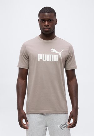 Homme portant un t-shirt beige Puma et un short gris clair, debout devant un fond blanc uni.